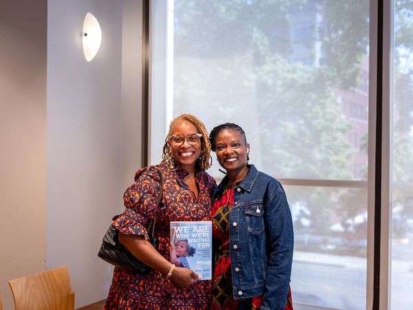 Two smiling women posing indoors, one holding a book titled 'We Are Who We're Waiting For'.