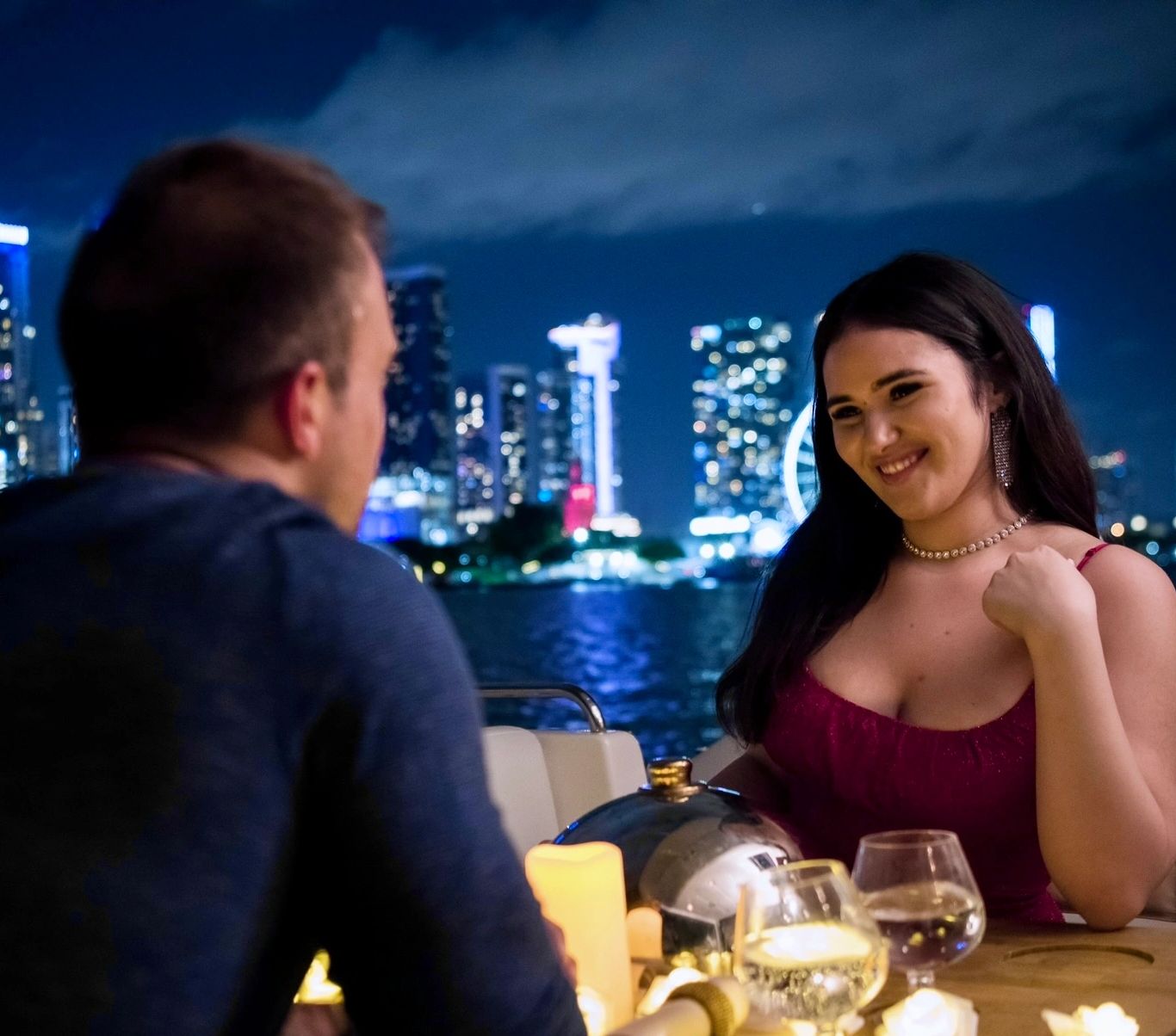 Couple enjoying a romantic dinner on a boat at night with city skyline in the background.