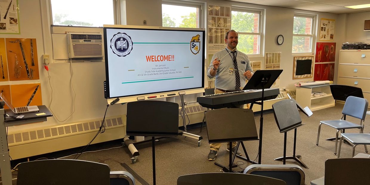 Music teacher welcomes students in a classroom with a keyboard and music stands.