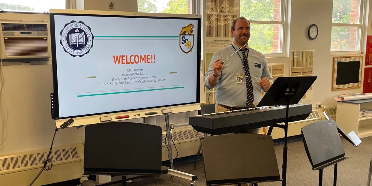 Music teacher welcomes students in a classroom with a keyboard and music stands.
