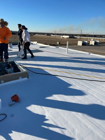Workers installing a white reflective roofing membrane on a flat roof under clear blue sky.