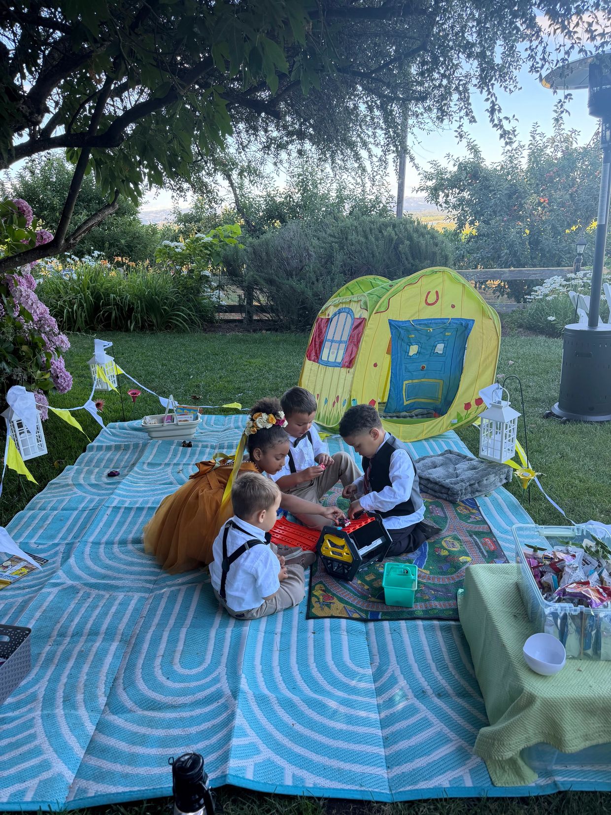 Children playing on a picnic blanket with a toy house in a garden.