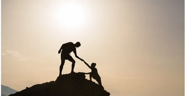 Two people on a mountain, one helping the other up at sunrise.