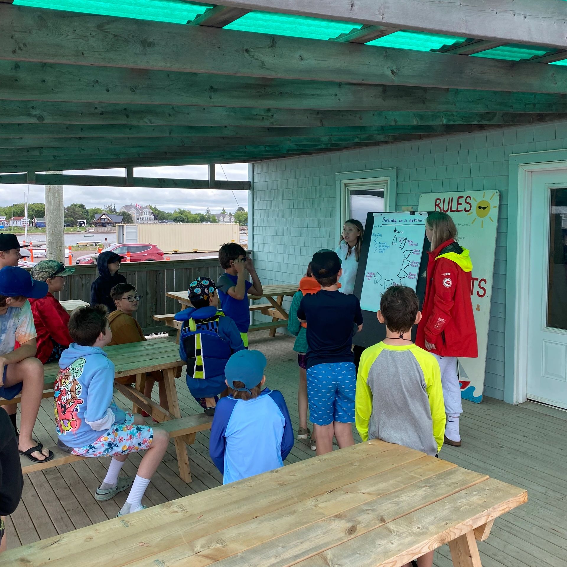 Group of children listening to a sailing lesson under a covered wooden patio.