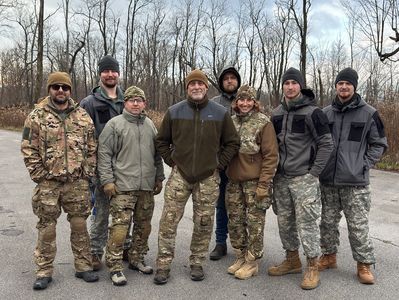 Group of eight people dressed in outdoor and camouflage clothing standing together outdoors.