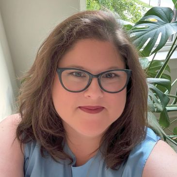 Woman with glasses smiling indoors near a window.