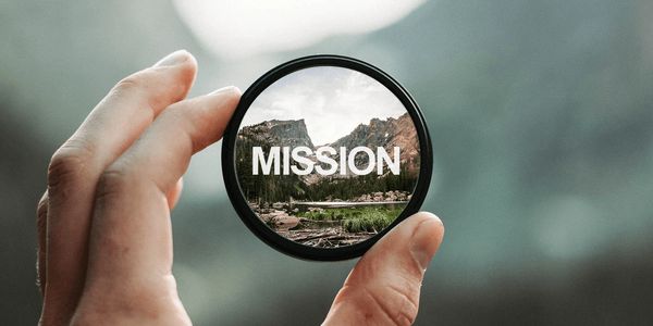 Hand holding a lens focusing on mountains with the word 'MISSION'.