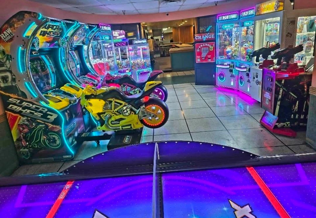 Arcade with neon-lit motorcycle racing and claw machines under bright lights.