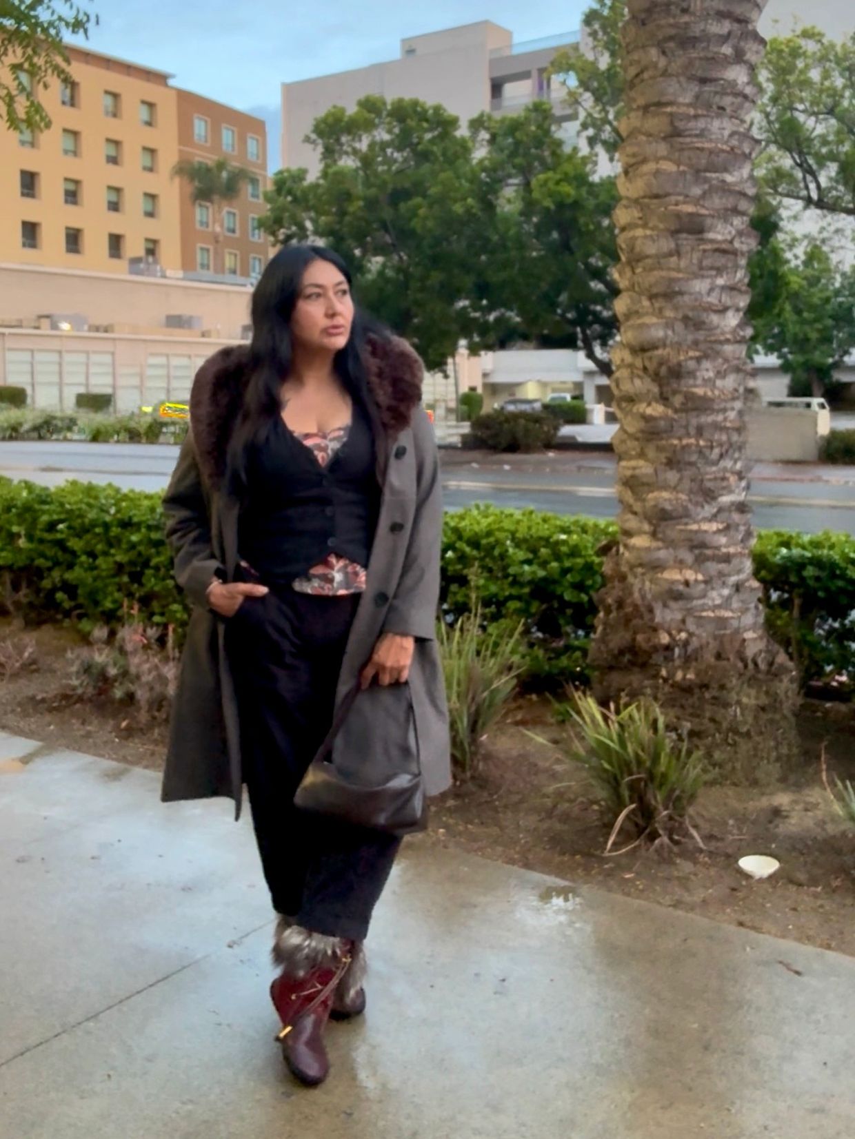 Woman in a fur-collared coat walking on a wet sidewalk near greenery and buildings.