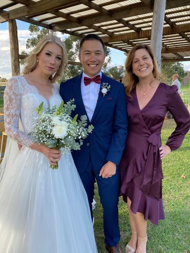 Bride, groom, and a guest posing outdoors under a pergola.