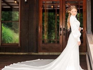 Bride in elegant white gown with long train standing on wooden porch.