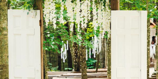 Rustic white doors open to a forest decorated with hanging white flowers and fairy lights.