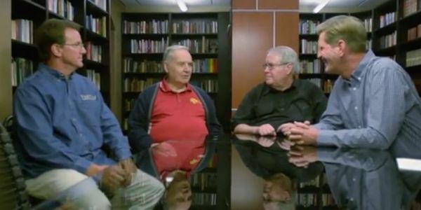 Four men engaged in a discussion around a glass table in a library setting.