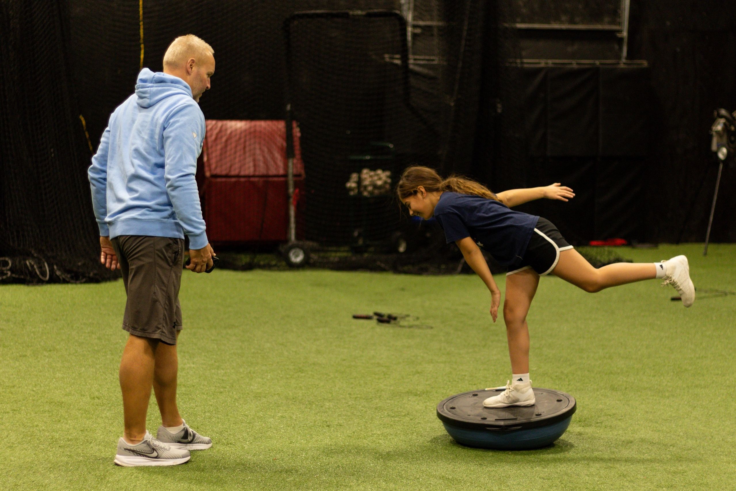 A girl balances on a Bosu ball under coach's guidance indoors.