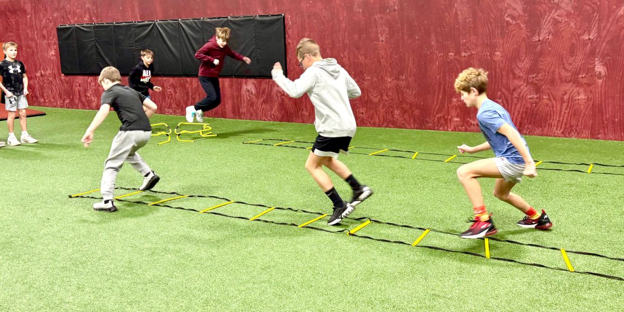 Kids doing agility drills with ladders and hurdles indoors on artificial turf.