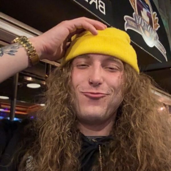 Person with long curly hair and yellow beanie smiling indoors.