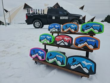 Colorful snowmobile goggles displayed on a snowy stand with a truck and tents in the background.