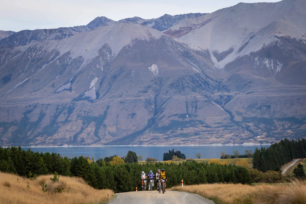 Cyclists riding on a gravel road with mountains and a lake in the background.