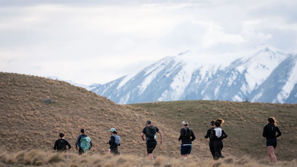 Group running on grassy terrain with snowy mountains in the background.
