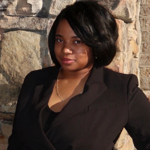 Woman in black blazer posing against a stone wall.
