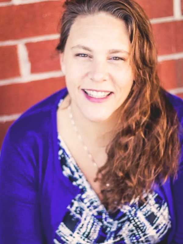 Smiling woman with long curly hair in a blue patterned dress and purple cardigan.