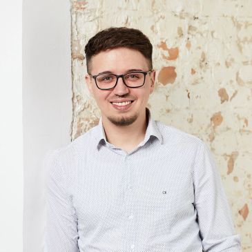 Junger Mann mit Brille und Hemd lächelt vor einer Wand.