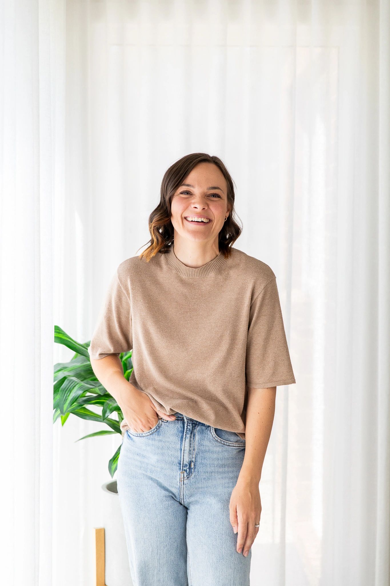 woman standing up and smiling, wearing a brown top and blue jeans with her right hand in her pocket