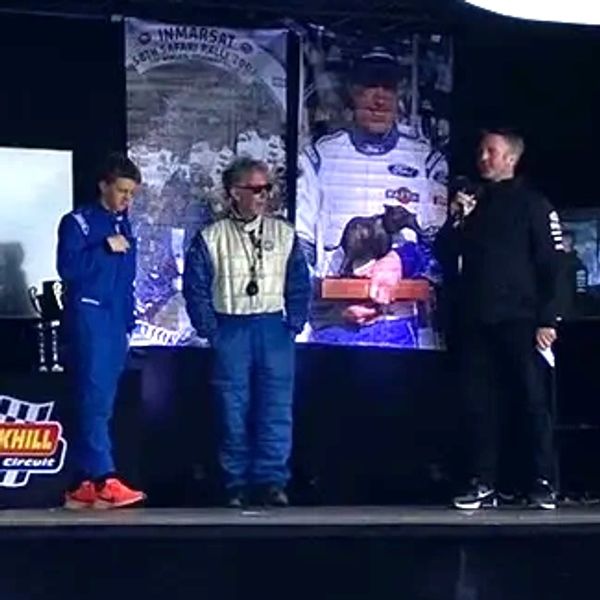 Three men on stage at a racing event with a large screen behind them.