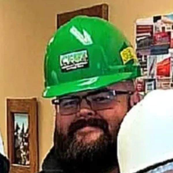 Man with beard wearing a green safety helmet and glasses.
