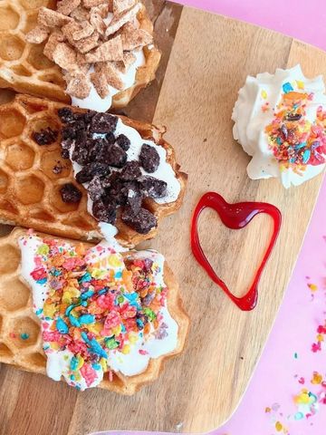 Waffles topped with whipped cream and colorful cereal on a wooden board with a heart-shaped syrup decoration.