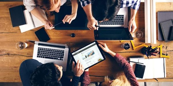 Four people collaborating over laptops and tablets at a wooden table.