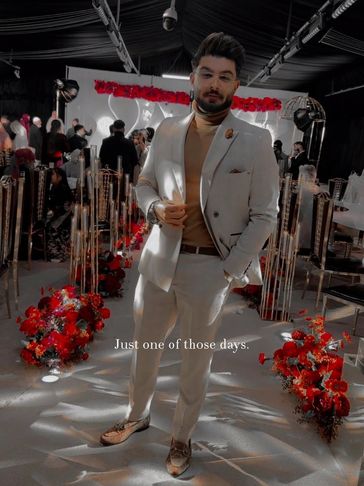 Man in beige suit posing at a decorated indoor event with red flowers.