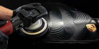 A person polishing a black Porsche car hood with a buffer.