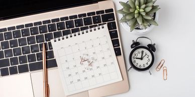 A notebook calendar placed on a keyboard next to a small alarm clock, paper clips and small plant