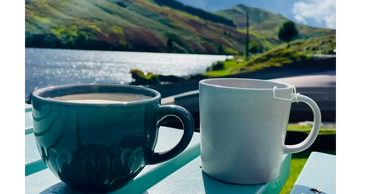 Coffee cups on table next to a lake on a sunny day. Colourful lush hills in background