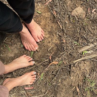 Two pairs of bare feet standing on dry, earthy ground with scattered twigs.
