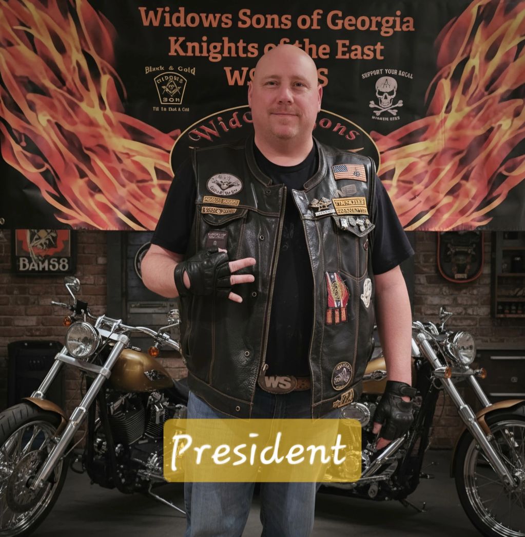 Man in leather vest standing confidently in front of motorcycles with 'President' label.
