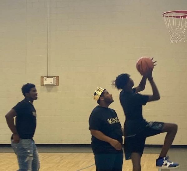 Three young men playing basketball indoors, one wearing a crown and a "KING" shirt.