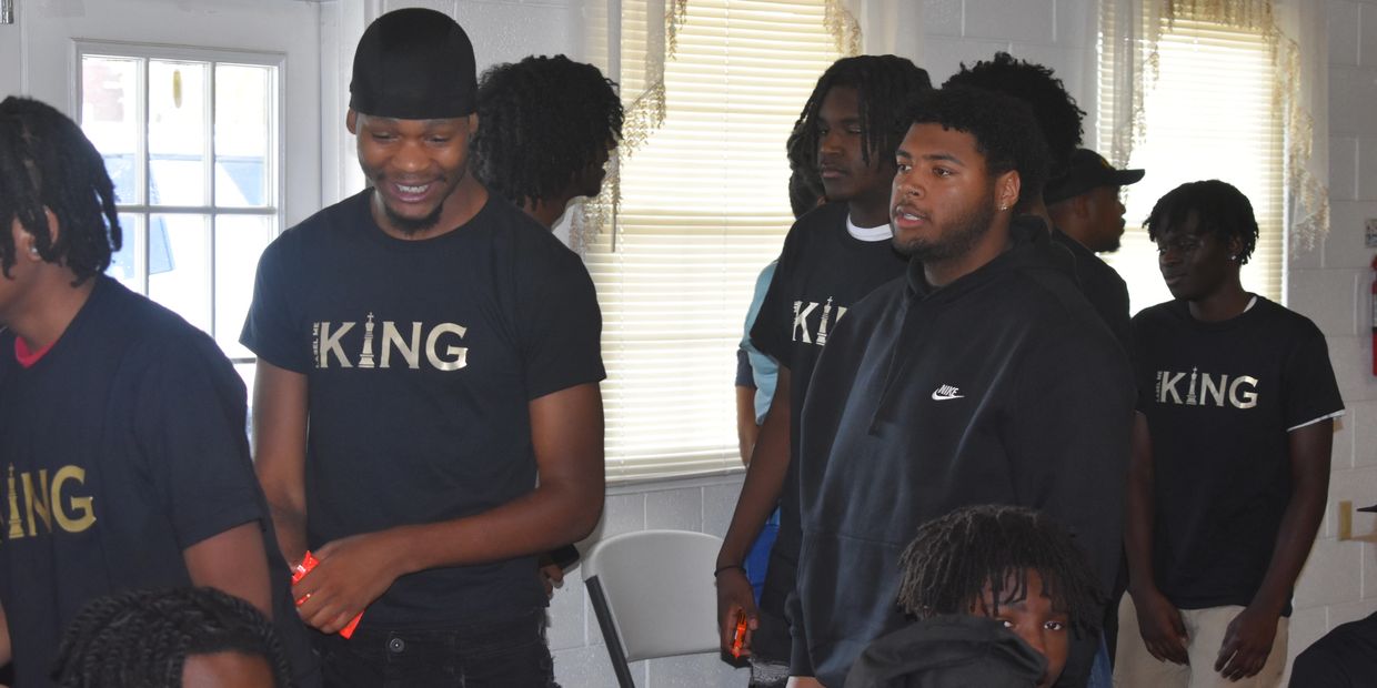 Young men gathered indoors, some wearing "KING" T-shirts, engaging in conversation.