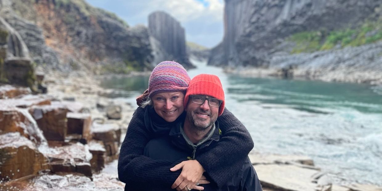 A couple embracing by a rocky river in winter hats.