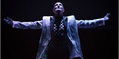 Performer in a checkered shirt and white jacket poses dramatically with arms outstretched.