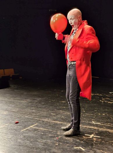 A clown in a red coat popping a red balloon on stage.