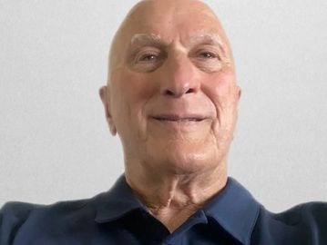 Smiling elderly man in a navy blue polo shirt against a plain background.