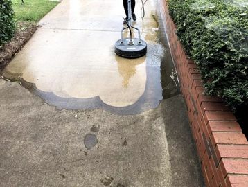 Person pressure washing a sidewalk near a brick wall and bushes.