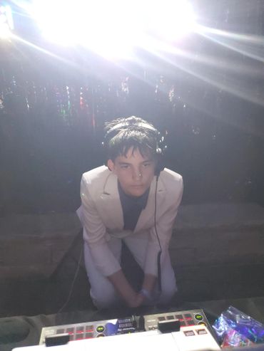 Young DJ in white suit performing at a dimly lit event with bright lights.