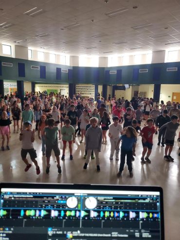 Large group of children dancing in a gymnasium with DJ equipment in foreground.