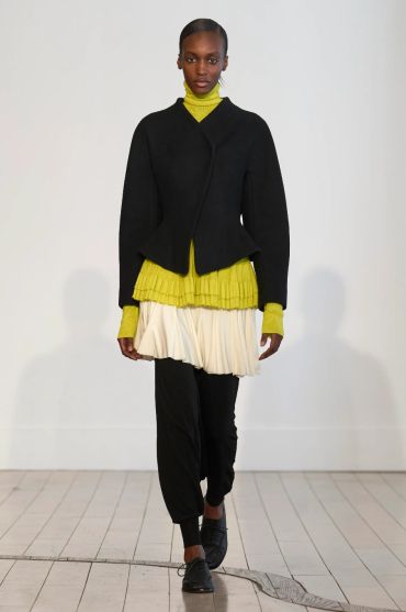 Model walking runway in layered outfit with black jacket and green top.