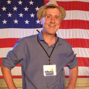 Man smiling in front of an American flag wearing a hat and badge.