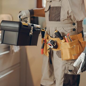 Handyman holding a toolbox while wearing a tool bet with various tools in a home setting.