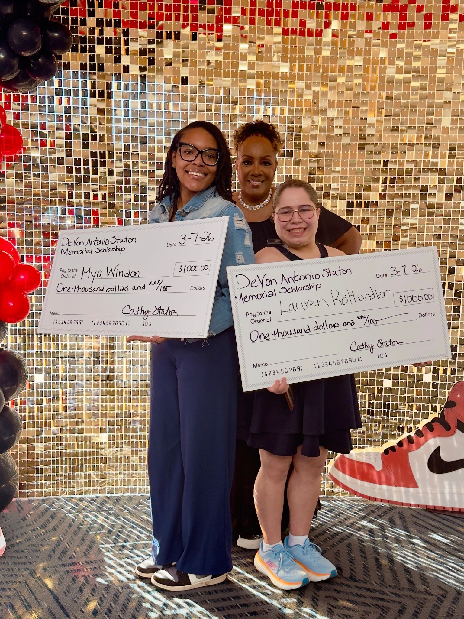 Two young women hold large scholarship checks, smiling with a woman behind them.